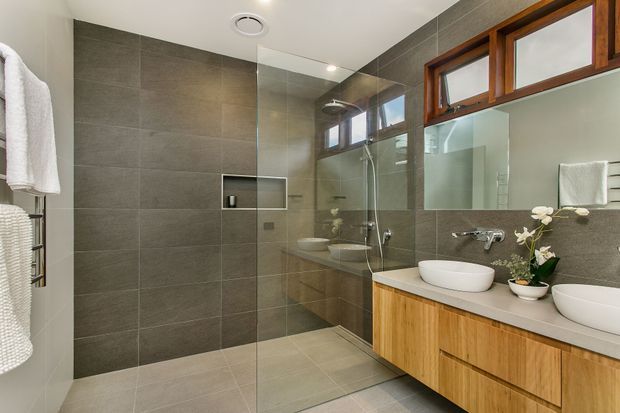 Contemporary bathroom with a large walk-in glass shower, grey stone-look wall and floor tiles, twin white vessel basins, and a long timber vanity with integrated drawers. Warm timber-framed windows, a mirrored wall, recessed shower niche, wall-mounted tapware, and simple decorative plants complete the space.