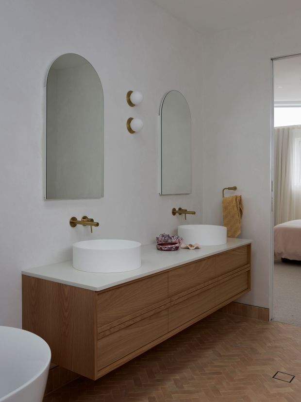 A refined contemporary bathroom featuring a long floating timber vanity with integrated drawers, a pale stone or solid-surface benchtop, two white vessel basins, wall-mounted brass tapware, and matching arched mirrors. Brass-and-white wall sconces, a brass towel rail, and warm herringbone flooring add detail. An open doorway reveals an adjacent bedroom.