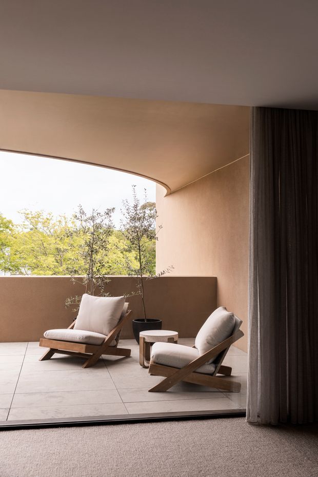 A refined indoor-outdoor lounge area viewed from inside, featuring two timber-framed lounge chairs with light upholstered cushions, a small round side table, a potted tree, and a warm beige rendered wall. Large curved glazing or an open corner connects the seating area to leafy greenery outside. Full-height dark curtains frame the opening, while large pale floor tiles transition to a textured carpeted interior floor. The space has soft natural light and a calm, warm, minimalist character.
