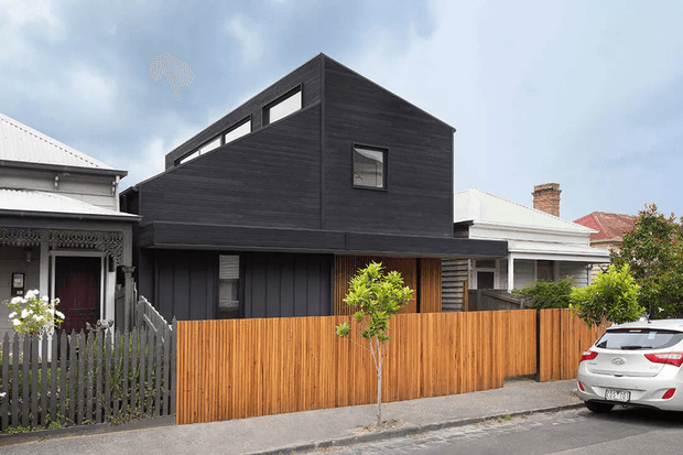 Street-facing view of a contemporary dark-clad multi-level house inserted between traditional neighboring residences. The home has a steep gable-like roof form, upper clerestory glazing, a projecting upper volume, timber screening and a warm vertical timber fence along the footpath.