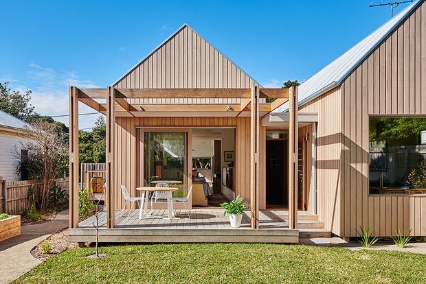 A bright exterior view of a contemporary timber-clad residential extension or compact house. The composition features vertical light-wood cladding, intersecting pitched roof forms, exposed timber posts and beams, a raised timber deck with outdoor dining furniture, large glazed doors, and a small landscaped lawn and garden. The open glazing provides a partial view into the interior, but the building exterior and deck dominate the image.
