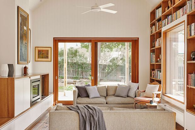 Bright, warm living room with a large upholstered sectional sofa facing a second sofa and a television or fireplace unit. Full-height timber-framed sliding doors open to a leafy outdoor area, while built-in timber bookshelves line the right wall. White vertical wall panelling, a ceiling fan, neutral upholstery, and rich timber joinery create a relaxed, refined interior.