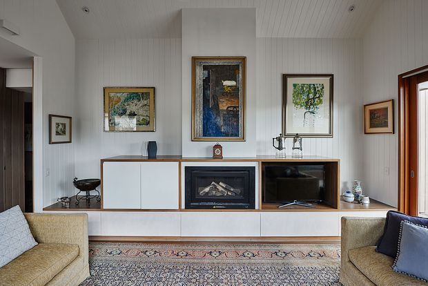 A bright, refined living room with white vertical wall panelling and a matching panelled ceiling. A built-in timber-trimmed media and storage wall incorporates a central fireplace, open shelving, and closed cabinetry. Framed artwork, a patterned area rug, and upholstered armchairs add warmth, while timber-framed glazing is visible at the right.