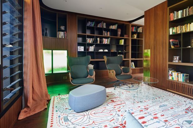 A richly detailed home library and reading lounge with extensive dark built-in shelving filled with books, warm vertical timber wall panelling, two teal upholstered lounge chairs, a pale oval ottoman, a glass coffee table, and a large patterned area rug. Floor-to-ceiling windows with sheer curtains bring in daylight, while green accent lighting adds a dramatic, sophisticated atmosphere.