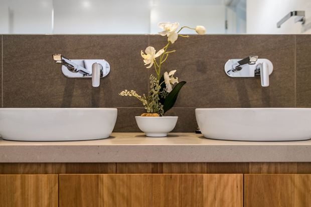 A close-up view of a refined double-vanity bathroom. Two white vessel basins sit on a pale stone benchtop above warm timber cabinetry, with wall-mounted chrome tapware and a dark brown tiled splashback. A small floral arrangement is positioned between the basins, and a mirror spans the upper wall.