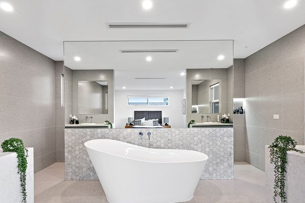 A bright contemporary bathroom featuring a freestanding white bathtub in the foreground, a double vanity with large mirrored wall, pale geometric patterned wall tiles, grey-beige porcelain wall and floor finishes, recessed ceiling lighting, and potted greenery. A bedroom is faintly visible through the central reflection/opening behind the vanity.