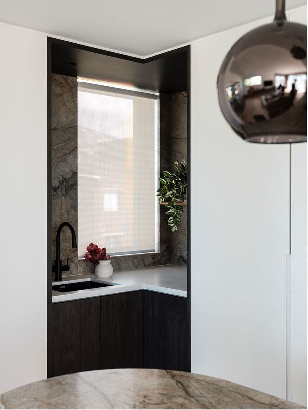 A compact, sophisticated kitchen corner viewed from an adjacent dining or living area. The kitchen features dark timber cabinetry, a pale stone benchtop, dramatic veined stone lining the window recess and splashback area, a black undermount sink with a curved black tap, and a roller blind over the window. A dark spherical pendant light and a marble-like foreground surface add a refined contemporary feel.