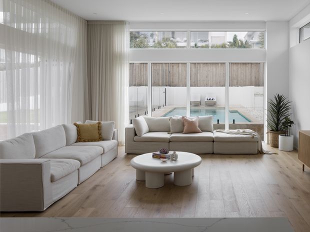 Bright, spacious contemporary living room with pale timber flooring, large floor-to-ceiling glazing, sheer curtains, and neutral upholstered sofas arranged around a rounded white coffee table. A private swimming pool and fenced courtyard are visible beyond the glazing. The room has a clean, calm, airy aesthetic with minimal built-in detailing and a light neutral palette.