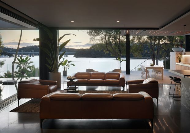 A sophisticated open-plan living room with a kitchen and dining area, framed by expansive floor-to-ceiling glazing overlooking a calm waterfront setting. The room features a large arrangement of cognac leather sofas and armchairs, dark polished flooring, a sculptural indoor plant, a compact dining table, and a sleek island kitchen with bar stools. The dark ceiling and minimal detailing create a refined, contemporary atmosphere.