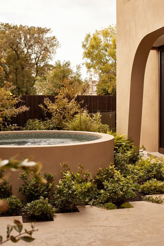 A warm, refined courtyard garden beside a stucco-clad building, featuring a curved raised plunge pool with a tiled interior, dense layered planting, a dark vertical timber privacy fence, and a large arched wall opening framing a dark glazed door. The paved courtyard and abundant greenery create an intimate, resort-like outdoor setting.