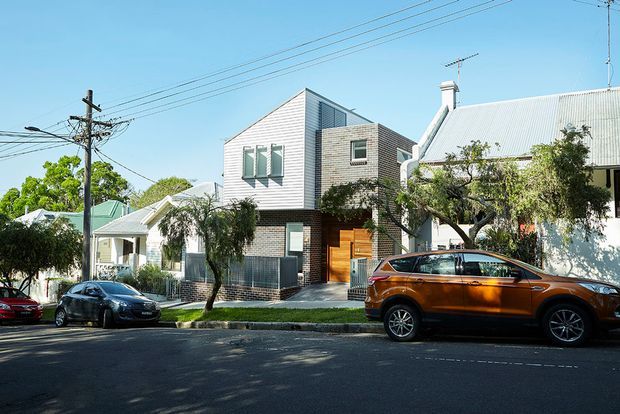 Street-level view of a contemporary multi-storey residence integrated into an established suburban streetscape. The home features a sharply pitched gabled roof, pale brick and light horizontal cladding, dark lower-level screening, timber entry doors, and prominent upper-level glazing. Adjacent traditional homes, mature street trees, overhead power lines, and parked vehicles are visible.