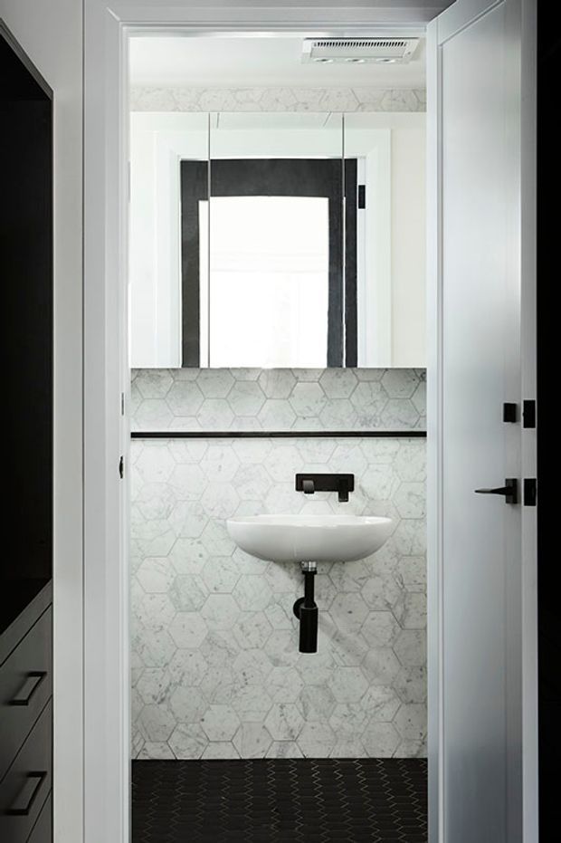 Compact contemporary bathroom viewed through an open doorway, featuring a wall-mounted white basin, matte black wall-mounted tapware and drain, mirrored cabinet, pale marble-look hexagonal wall tiles, and dark patterned floor tiles. White trim and cabinetry create a crisp, high-contrast scheme.