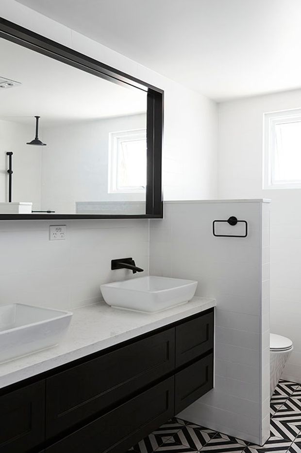 Bright contemporary bathroom with a double vanity, white vessel basins, dark built-in cabinetry, white stone-look benchtops, black tapware and mirror frames, and bold black-and-white geometric floor tiles. A toilet is visible behind a partial tiled privacy wall.