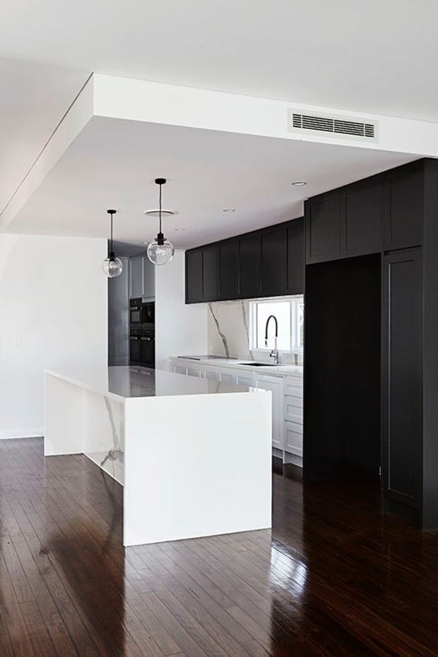 A contemporary kitchen with a compact white island, dark charcoal-black full-height cabinetry, white marble-look surfaces with grey veining, integrated appliances, and a contrasting dark timber floor. Clear globe pendant lights hang above the island, while a large dark pantry or storage wall dominates the right side.