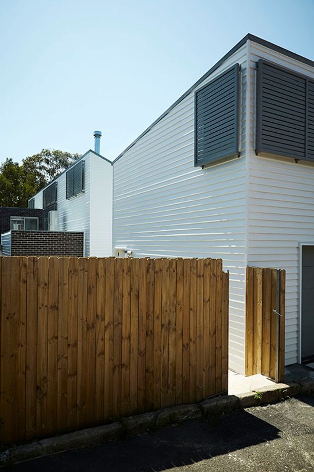 Exterior view of a compact contemporary residential building clad in white horizontal weatherboards. The facade features dark louvred window shutters, a small chimney, a dark garage or service door, and a timber privacy fence along the street edge.