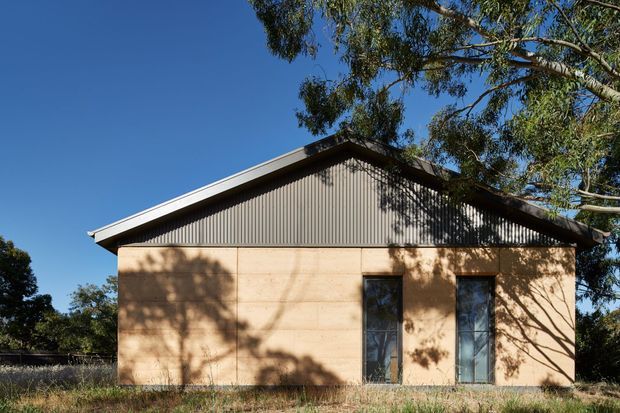 Exterior view of a compact single-storey building with a simple gable roof, corrugated metal upper facade, warm tan rendered or panelled lower walls, and narrow vertical windows. Set among trees under a clear blue sky.