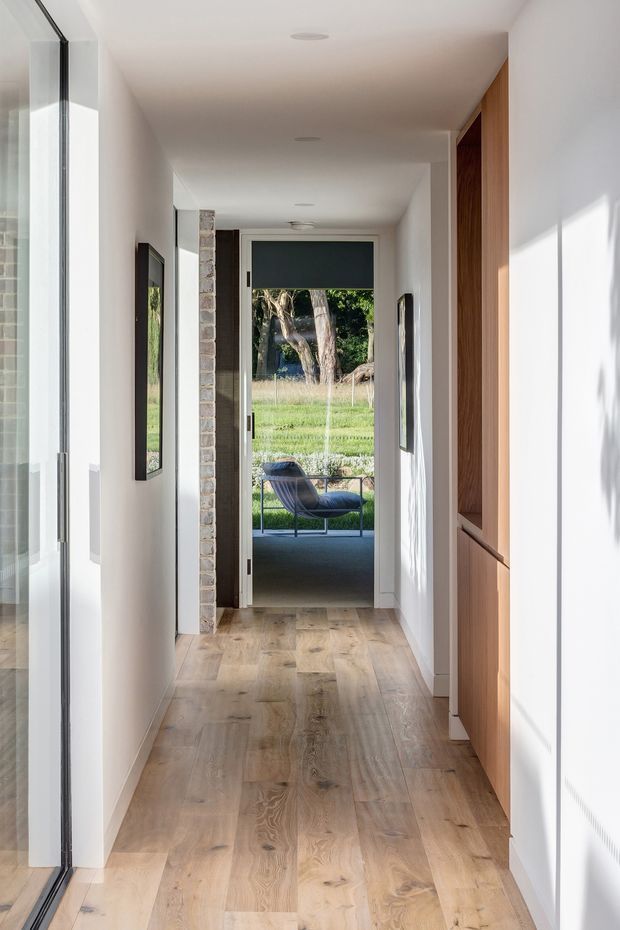 A bright, narrow residential hallway leading to a glazed exterior door and outdoor seating area. The corridor has white painted walls and ceiling, recessed downlights, warm timber flooring, a full-height timber built-in storage unit on the right, framed artwork, and a partially exposed brick structural or feature wall near the doorway. Strong daylight enters through full-height glazing on the left and the rear door, creating a clean, calm connection to the garden beyond.