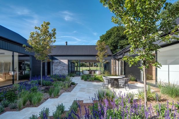 A landscaped courtyard surrounded by a contemporary single-storey building with dark corrugated metal roofing and cladding, large glazed openings, and exposed light brick walls. Pale stone paving forms a central walkway and patio with outdoor dining furniture, while structured planting and purple flowering groundcover frame the space. The courtyard is bright, airy, and carefully integrated with the surrounding architecture.