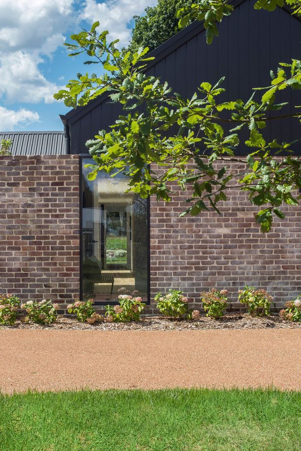 Exterior view of a contemporary building with a dark vertically clad gabled upper volume above a low red-brown brick wall. A tall glazed doorway or window provides a view into the building, while a landscaped garden bed, gravel path and lawn sit in the foreground. Mature leafy planting partially screens the facade.