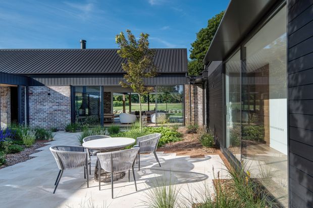 A contemporary courtyard viewed from outside, with a paved outdoor dining area furnished with a round table and four chairs. The courtyard is enclosed by dark horizontal cladding, pale brick walls, extensive glazing, and a low corrugated metal roof. Native-style planting softens the edges, while the glazed living spaces and a sheltered seating area are visible beyond.