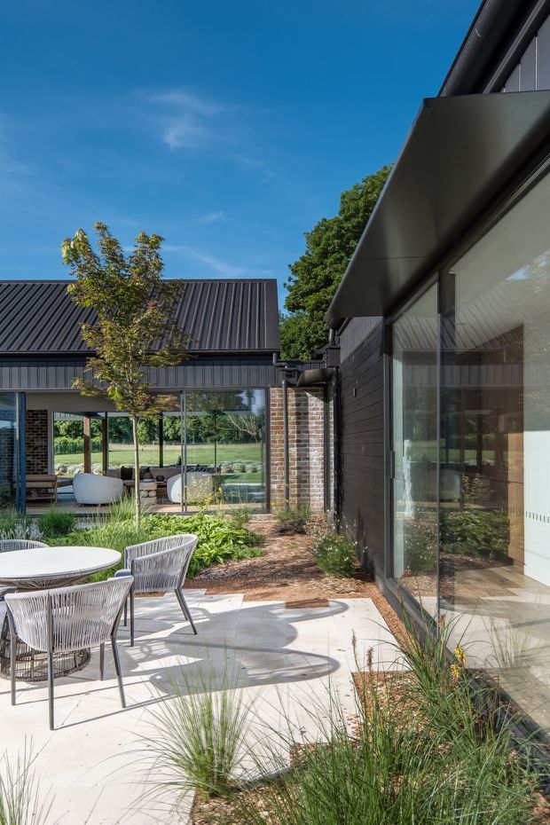 A landscaped courtyard patio beside a contemporary dark-clad house, with pale concrete paving, outdoor dining furniture, ornamental grasses and garden planting. Large glazed openings connect to a modern living space, while a brick-clad link and corrugated metal roof define the surrounding architecture. The view looks toward an open green rural or suburban landscape under a clear blue sky.
