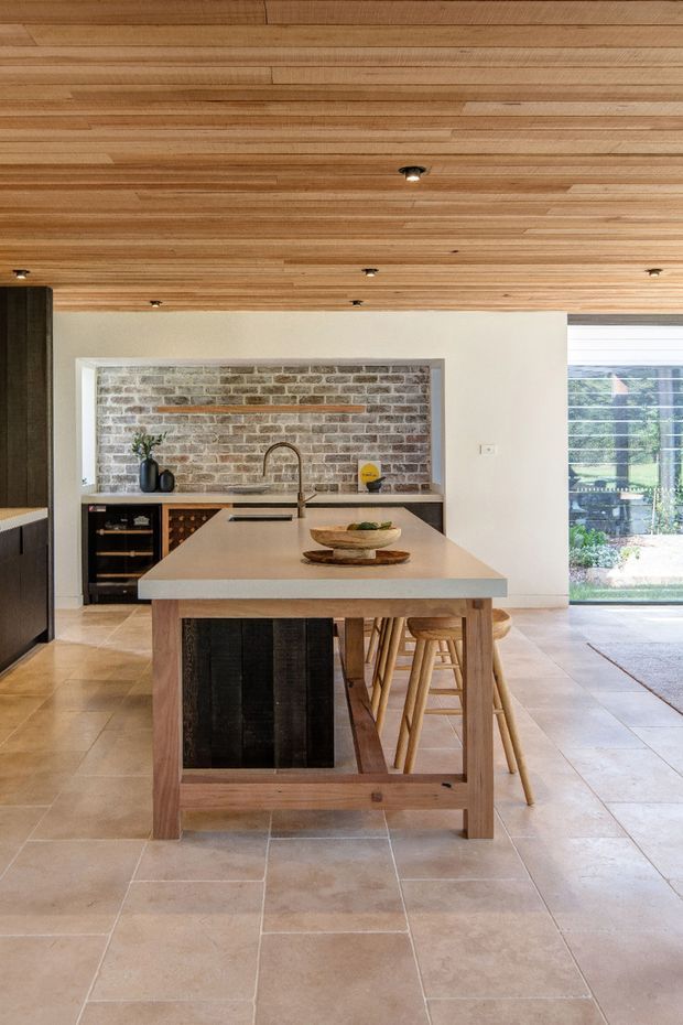 Bright contemporary kitchen viewed toward a large central island. The room features a timber-lined ceiling, pale natural stone or porcelain floor tiles, a light stone island benchtop, timber island framing with dark vertical panel detailing, and a brick feature splashback behind the sink. Timber bar stools sit beneath the island, with large glazed doors or windows opening to greenery outside.