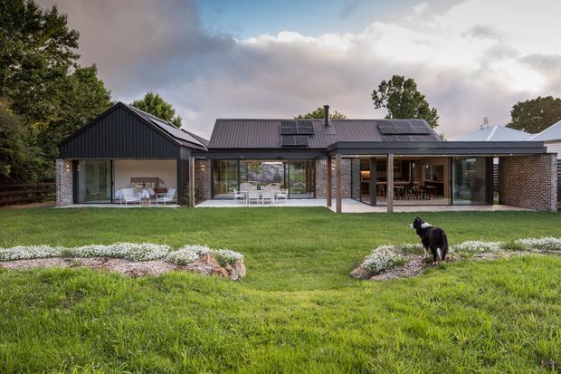 Wide exterior view of a low-profile contemporary home set in a landscaped lawn. The house features dark vertical metal cladding, exposed brick walls, extensive floor-to-ceiling glazing, multiple pitched roof sections with skylights or solar panels, and covered outdoor living areas. Garden beds and a dog occupy the foreground.