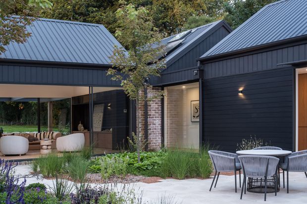 Contemporary dark-clad residential courtyard exterior with multiple pitched metal roof forms, glazed living spaces, a brick accent wall, landscaped garden beds, and an outdoor dining setting. The composition highlights the building facade and sheltered outdoor living area.