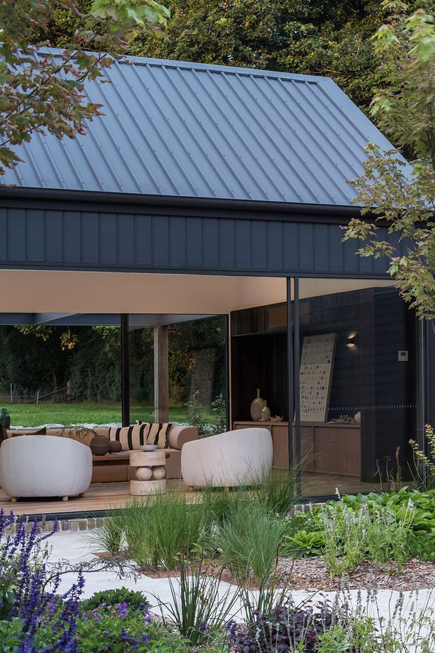 A contemporary residential exterior viewed from a landscaped garden. The house features a steep corrugated metal gable roof, dark vertical cladding, large floor-to-ceiling glazed openings, and a sheltered outdoor living area furnished with lounge seating. Layered ornamental planting and pale garden paving fill the foreground.