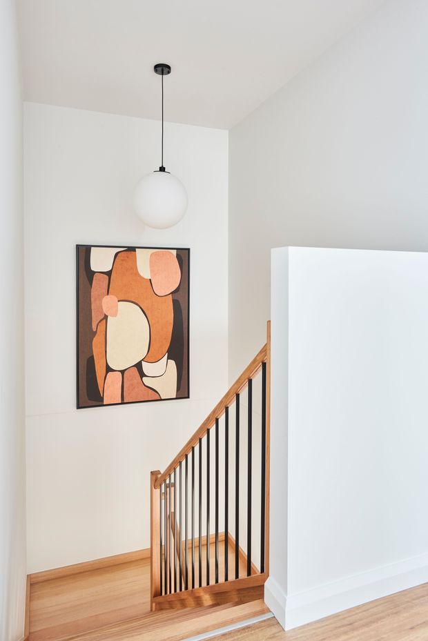 A bright, minimalist stair landing with white walls and ceiling, warm timber flooring and stair handrail, and black vertical metal balusters. A spherical pendant light hangs above an abstract framed artwork in warm terracotta, cream, and brown tones. The stairwell is partially screened by a clean white wall volume.