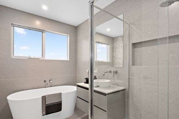 Bright contemporary bathroom featuring a freestanding white bathtub, wall-mounted brushed-metal tapware, a floating vanity with a white vessel basin, large-format grey wall and floor tiles, and a frameless glass shower enclosure with a recessed niche. A wide window and mirror reflect the clean, airy space.