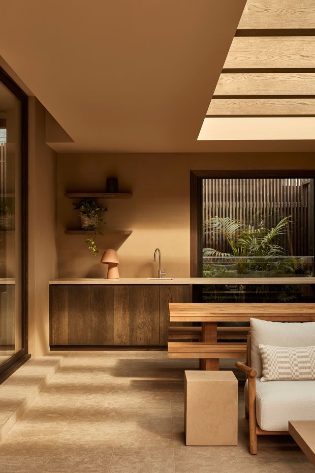 Warm, refined interior showing a compact open-plan living and kitchenette area. The room features dark timber cabinetry with an integrated sink, earthy plaster walls, large stone-look floor tiles, timber-framed glazing, floating timber shelves, and a skylight with exposed timber slats. A timber dining table and upholstered lounge chair occupy the foreground, while plants are visible through the window and on the shelves.