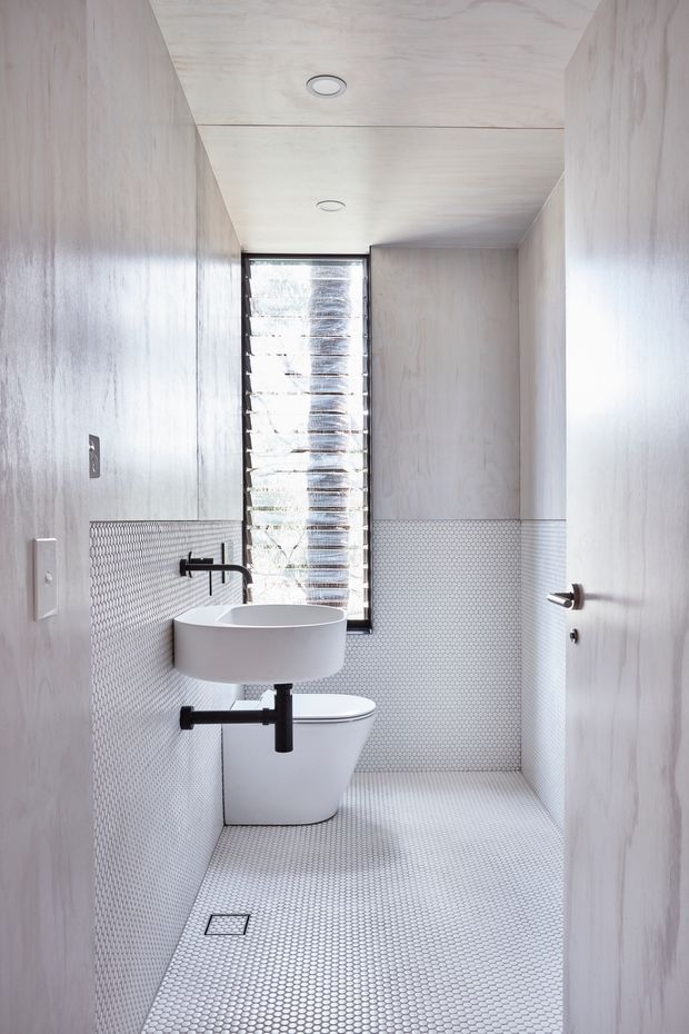 A narrow, compact bathroom with pale timber-lined walls and ceiling, white hexagonal mosaic tiles covering the lower walls and floor, and a wall-mounted white basin and toilet. Matte black tapware, flush plate, and door hardware provide contrast. A tall louvred window brings in natural light at the end of the room, with recessed ceiling lights above.