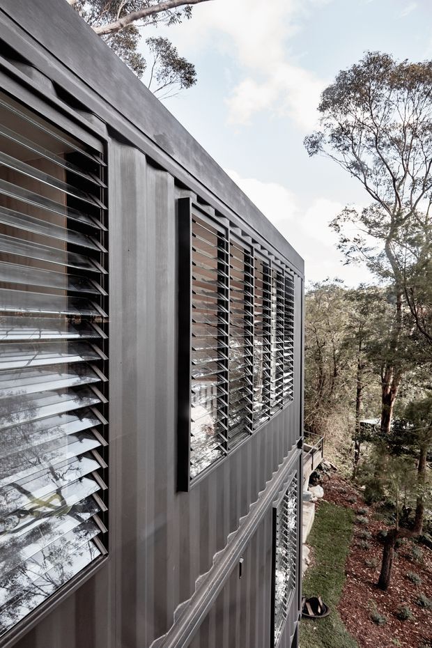 Close-up exterior view of a contemporary multi-level building set among trees, featuring dark grey corrugated cladding and repeated horizontal louvred window screens. The elevated facade overlooks a wooded, sloping site and emphasizes passive shading and ventilation elements.