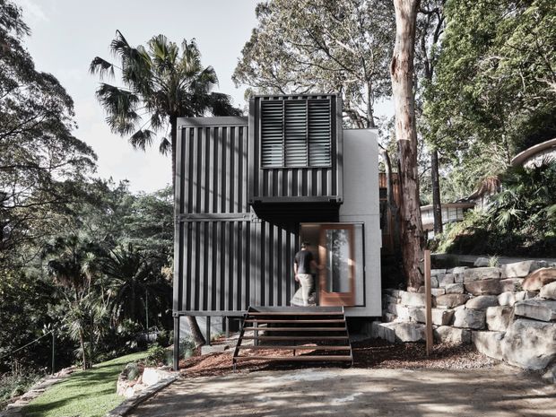 A compact modern dwelling set into a densely wooded, sloping site. The dark vertically ribbed facade includes projecting box-like volumes, horizontal louvres, and a glazed entry, with a small timber stair leading up from a paved driveway. Stone retaining walls and mature trees surround the building.