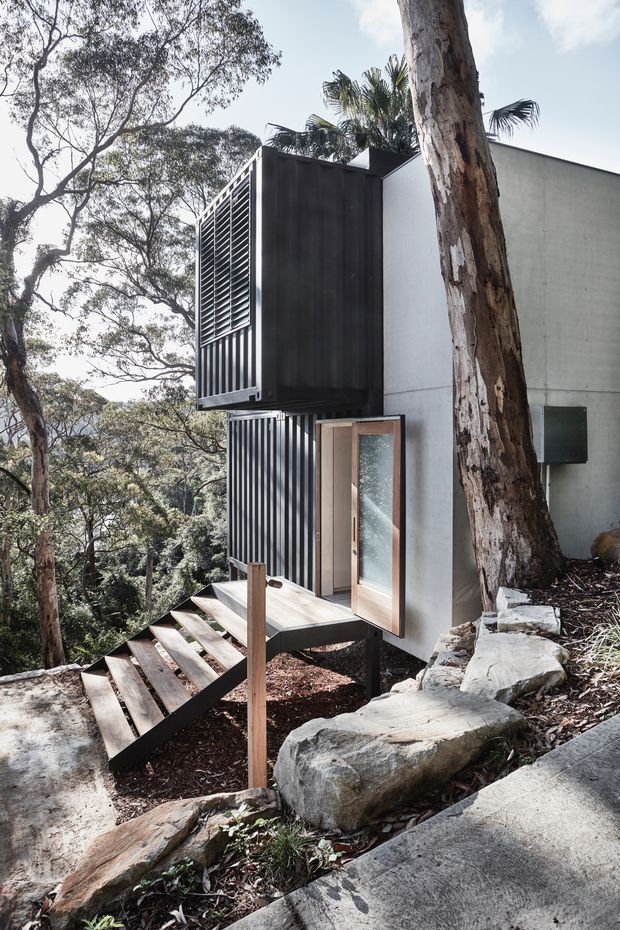 A compact modern house set into a steep, wooded site. The exterior combines smooth pale walls with dark vertical metal cladding and louvred screening, while a timber-framed glazed door opens onto a small elevated timber landing and stair. Large natural rocks, eucalyptus trees and dense bush surround the structure, emphasising its sensitive hillside setting.