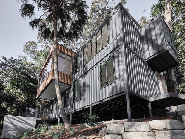 Exterior view of a multi-level contemporary house built with strongly articulated corrugated metal cladding, elevated on dark structural posts above a rocky sloped site. The building has projecting box-like volumes, louvred openings and balconies, with warm timber-clad sections contrasting against the grey metal envelope. Dense vegetation and palm trees surround the house.