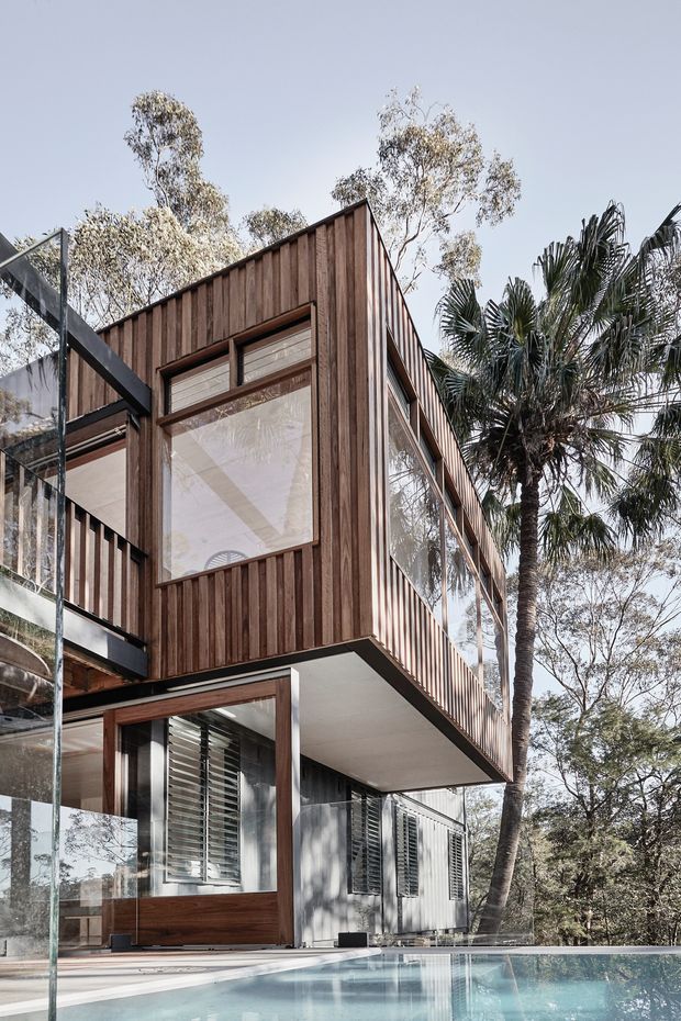 A contemporary multi-level house exterior beside a swimming pool, viewed from a low angle. The upper storey projects over the lower level and is clad in vertical dark timber boards, with extensive floor-to-ceiling glazing and slim dark frames. The lower level combines pale rendered walls, timber-framed openings and external louvres. Glass balustrades, a cantilevered upper volume and surrounding palms and mature trees contribute to a refined, warm modern residential setting.