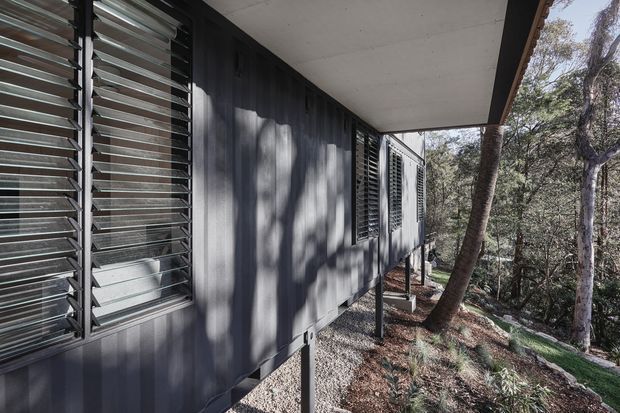 Exterior side view of a dark-clad elevated building set among bushland. The facade features extensive horizontal louvred windows for ventilation and sun control, with a simple projecting roof canopy and slender supports beneath the structure.