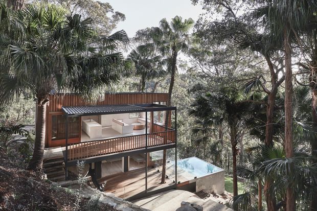 A multi-level contemporary house nestled within dense tropical vegetation on a sloping site. The home features timber-clad walls, broad outdoor decks with dark metal framing and balustrades, extensive glazing, and a small swimming pool on the lower terrace. The elevated, tree-surrounded composition creates a strong connection to the natural landscape.