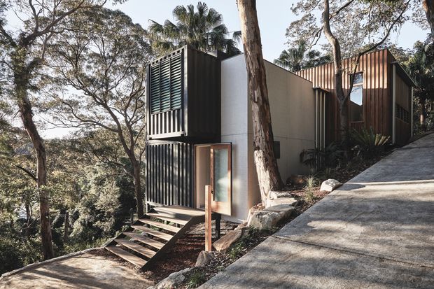 Exterior view of a compact contemporary multi-level dwelling set into a steep, wooded site. The building combines dark vertical cladding, pale rendered walls, extensive louvred screens, and a pitched roof form. Timber steps lead to a glazed entry, while a sloped concrete driveway occupies the foreground. Mature trees and dense vegetation surround the structure.