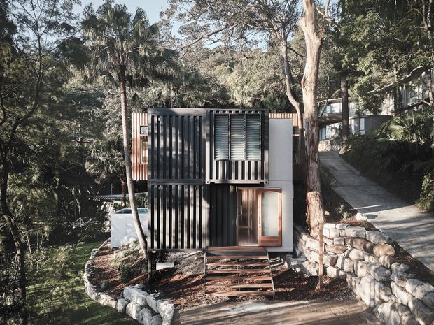 A compact modern dwelling set into a heavily wooded, sloping site. The dark corrugated metal-clad facade features strong vertical ribbing, projecting window surrounds with horizontal louvres, and a small timber-framed entry door. Stepped timber decking and stone retaining walls lead up to the building, while a larger structure is partially visible through the trees behind it.