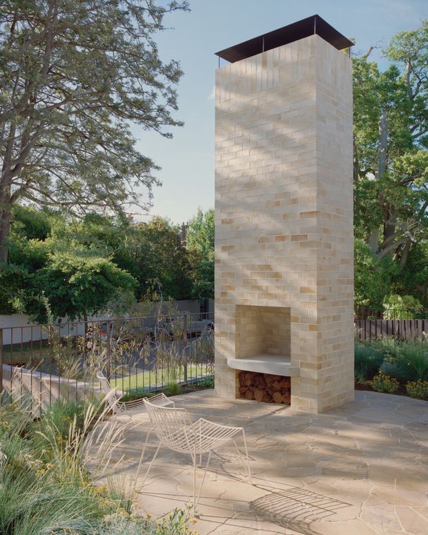 A landscaped outdoor courtyard or terrace featuring a tall, freestanding masonry fireplace with a broad dark metal cap. The fireplace is built from pale beige rectangular blocks and includes an open firebox with stacked firewood below. Paved stone surrounds the structure, with wire-frame lounge chairs, ornamental grasses, shrubs, mature trees, and a simple metal railing creating a relaxed garden setting.