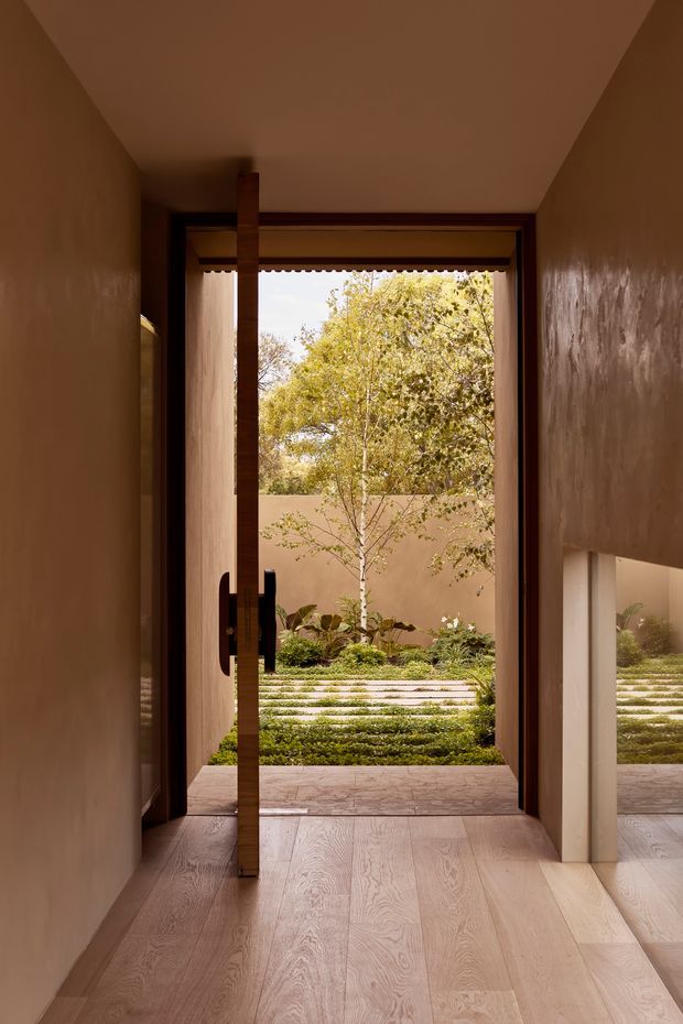 A warm, narrow interior entryway or hallway leads to a full-height glazed timber door opening onto a landscaped courtyard. The space has smooth earthy plaster walls, pale timber flooring, dark timber door framing, and a large reflective panel on the right. The courtyard beyond contains layered planting and horizontal stepping or paving bands, with mature trees visible outside.