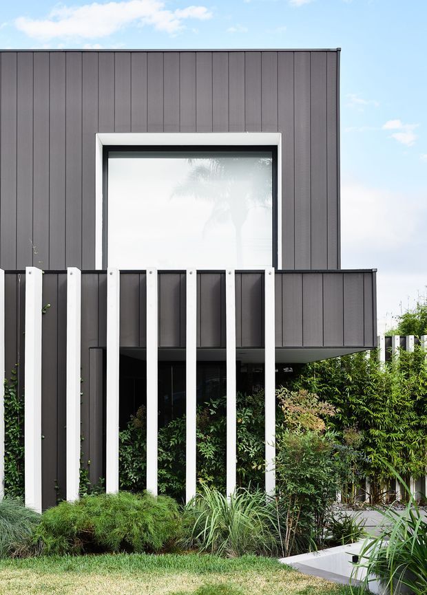 Exterior view of a contemporary two-storey house with dark vertically ribbed cladding, a large projecting upper-level window, and a projecting balcony or box form. White vertical structural or screen elements frame the lower facade, which is partially concealed by dense landscaped planting and grasses.