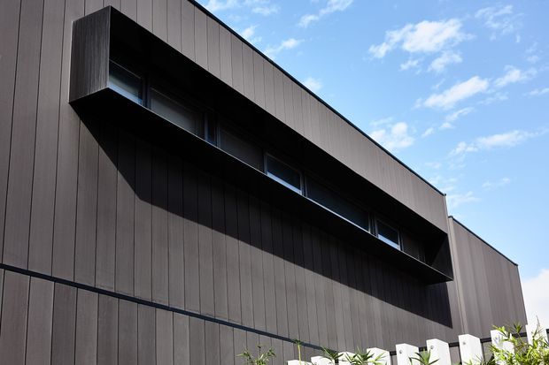 Close-up exterior view of a contemporary, rectilinear building facade clad in dark vertical panels. A deep projecting upper volume creates a dramatic shadow over a continuous strip of narrow windows, with a small section of white fencing and planting visible at the base against a bright sky.