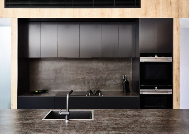 A contemporary kitchen with a dark matte-black built-in cabinetry wall, integrated double ovens, black cooktop, and dark textured backsplash. A broad dark stone-look island with an undermount sink and sculptural black tap occupies the foreground, framed by pale timber wall panels and a black linear ceiling vent.