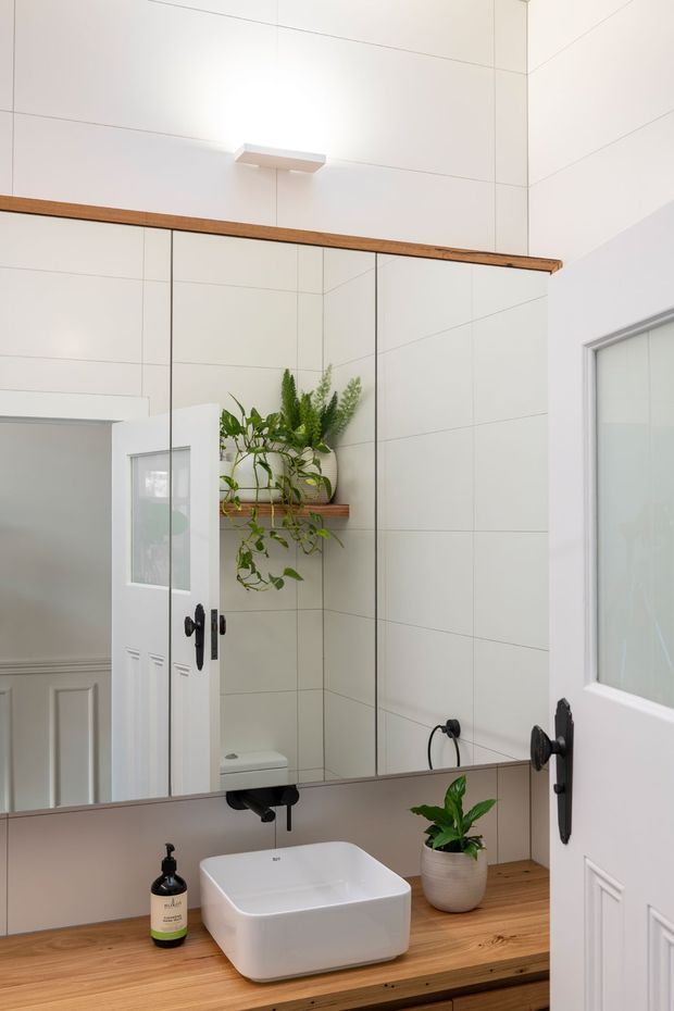 Bright, compact bathroom vanity with a white vessel basin set on a warm timber benchtop, black wall-mounted tapware, and a large mirrored wall. White tiled walls, timber shelving with potted plants, and a partially visible toilet and doorway create a clean, contemporary look.