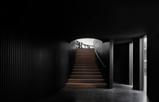 A dramatic, dark stairway interior with broad timber-toned steps rising toward a bright upper level. The walls and ceiling are predominantly black, with vertically ribbed wall panelling on the left, dark structural partitions, slim linear handrails, and narrow vertical windows on the right. The image has a highly atmospheric, sculptural and monochromatic appearance.