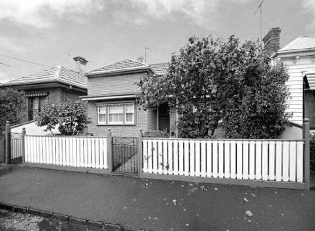 Black-and-white streetscape photograph of a modest single-storey residential dwelling, likely a period worker's cottage or bungalow, behind a white picket fence. The house has a tiled hipped roof, brick walls, timber-framed windows, a small front garden with mature shrubs, and a paved street frontage. A neighbouring weatherboard house is partially visible at right.
